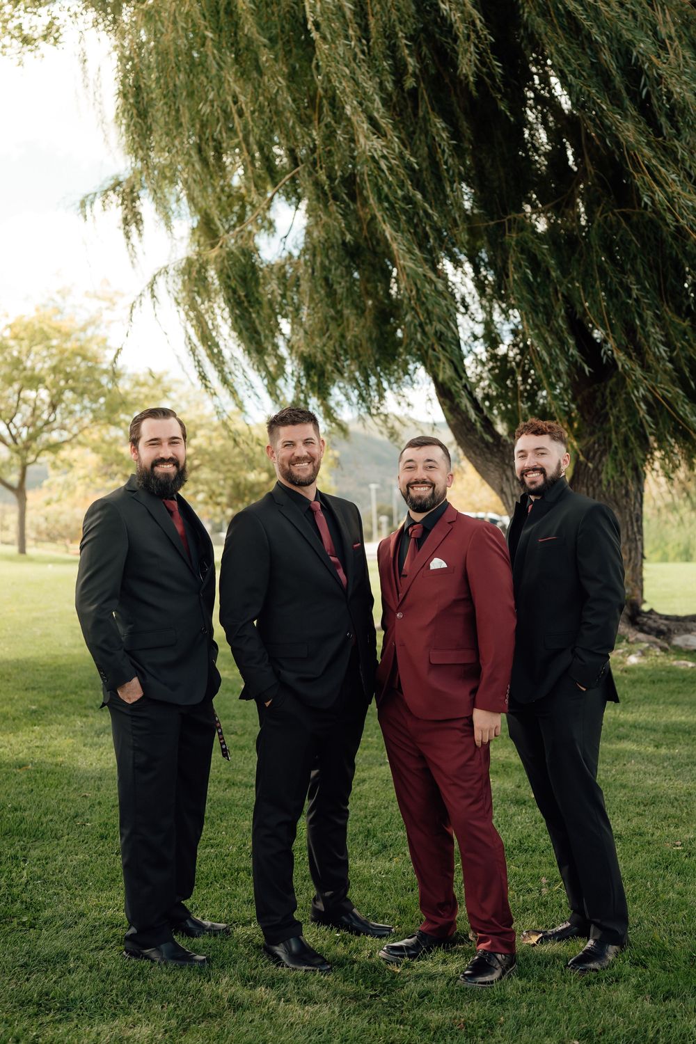 Four groomsmen in black and burgundy suits pose together on grassy lawn under willow tree.