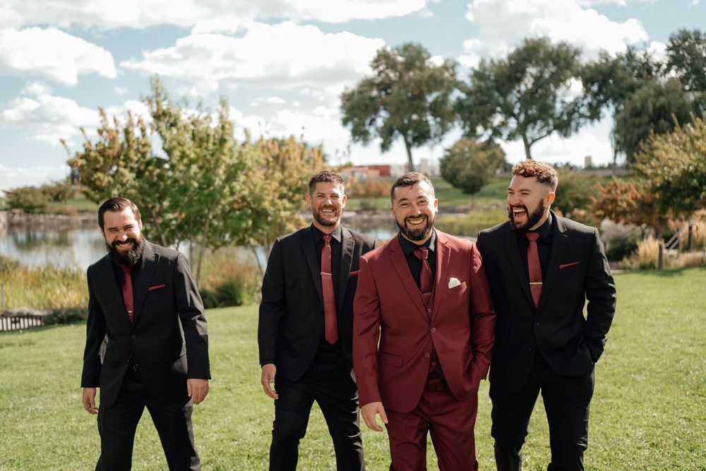 Groomsmen in burgundy suits and black jackets laugh together while walking on grass in front of trees.