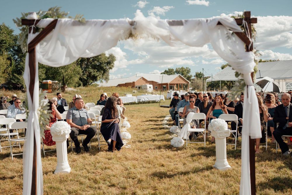 An outdoor wedding ceremony setup with white fabric draped arches and chairs on a grassy field under blue skies.