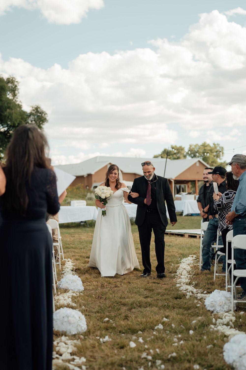 Wedding ceremony processional on a grassy field with white floral decorations and guests watching.