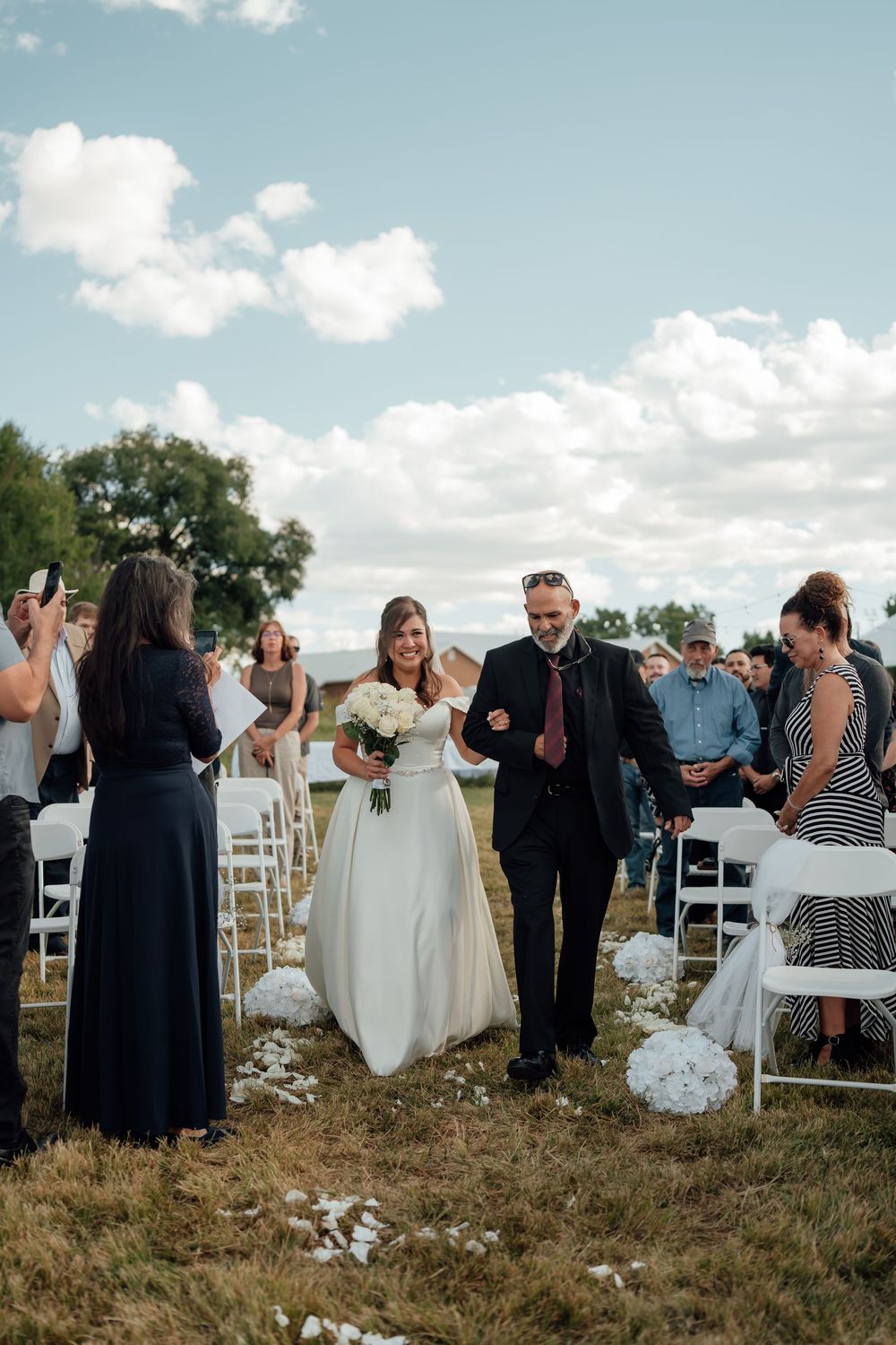 Wedding guests gather on a grassy hillside with white chairs for an outdoor ceremony.