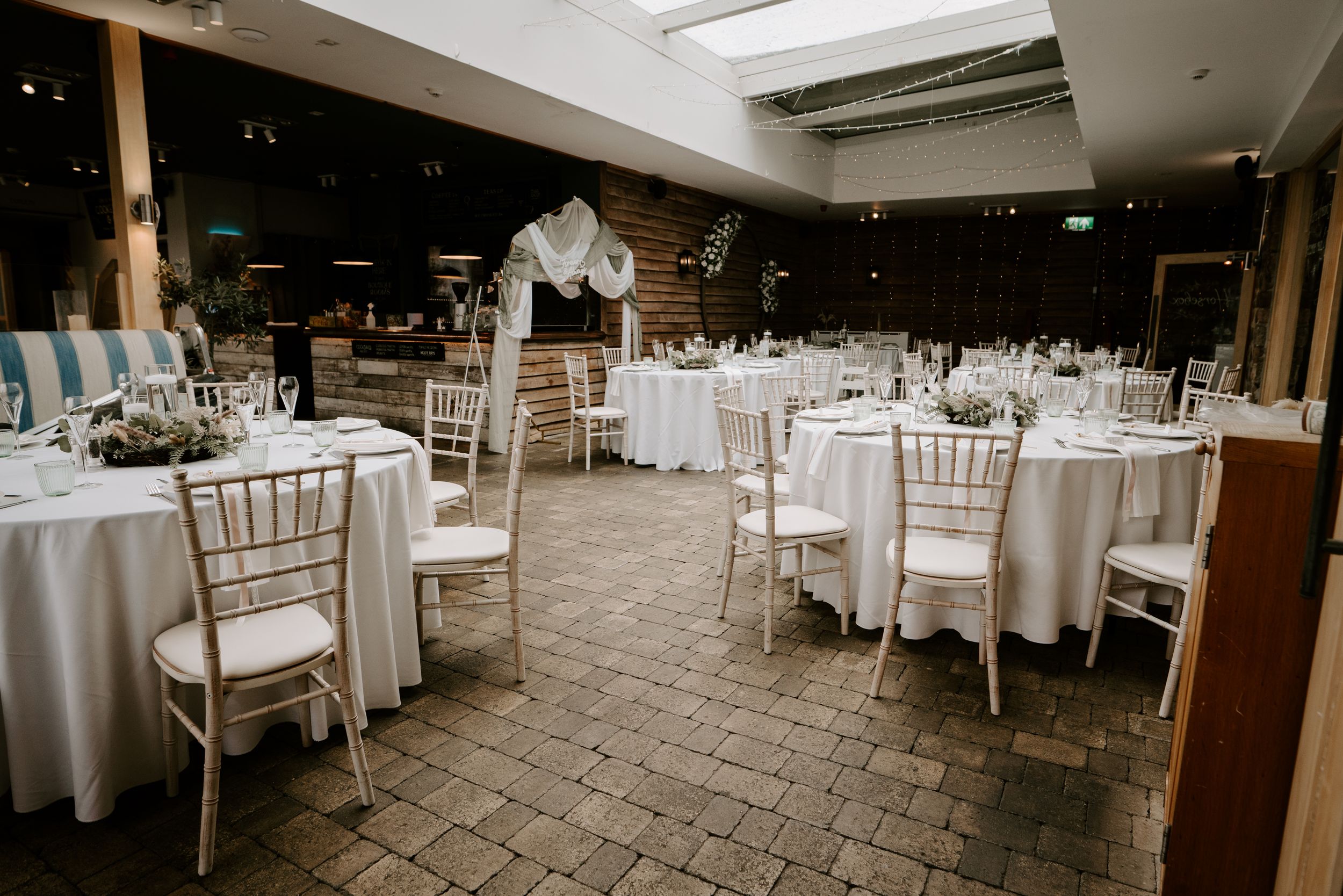 An elegant wedding reception venue with white round tables, chiavari chairs, and a brick floor under skylights.