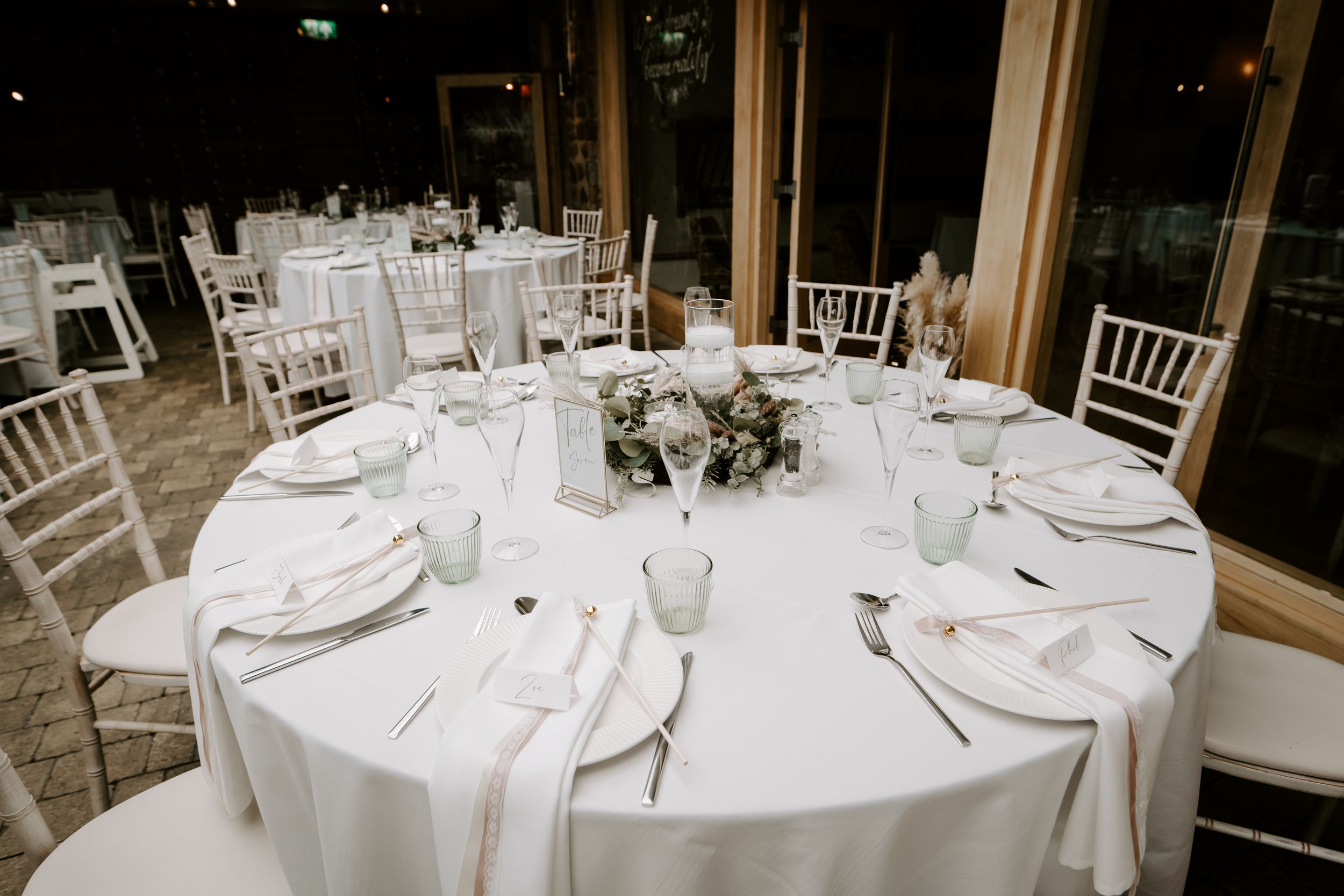 Round tables with white linens and elegant place settings arranged in a formal dining room with natural lighting.