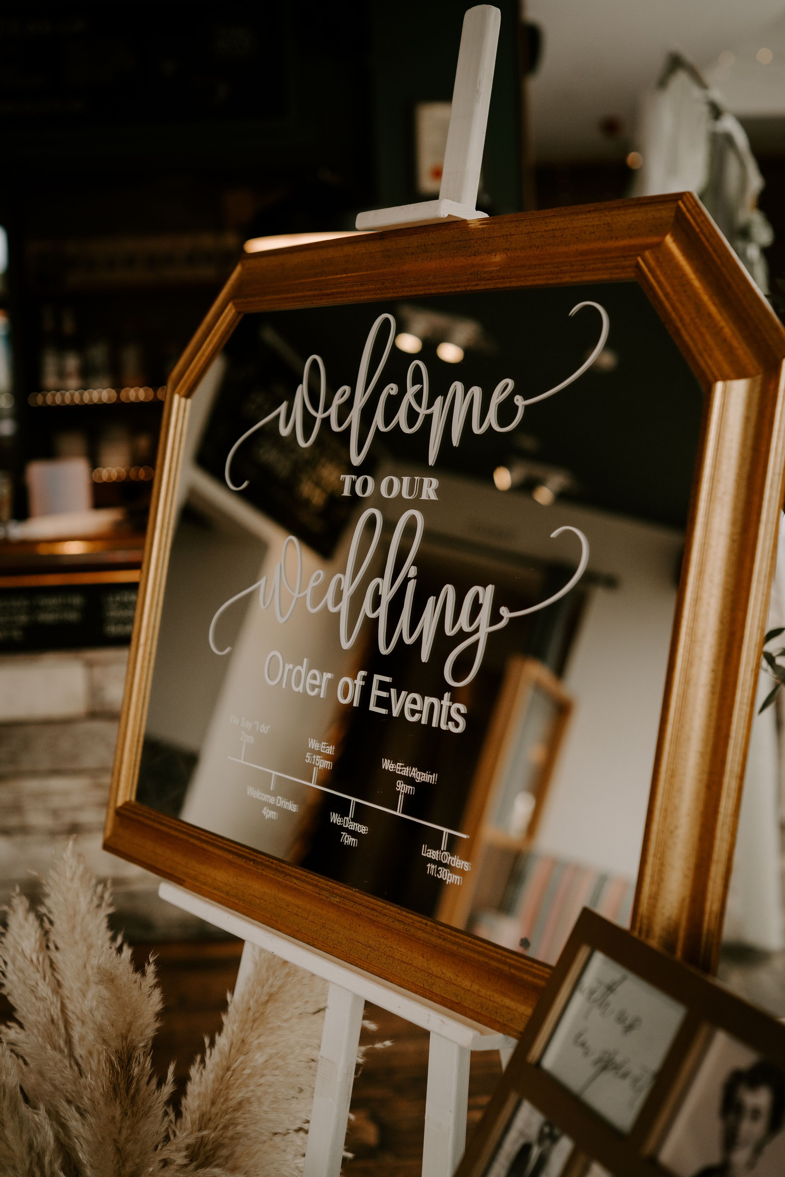 Rustic wooden framed wedding signage displays welcome messages and event details with dried pampas grass decorations.