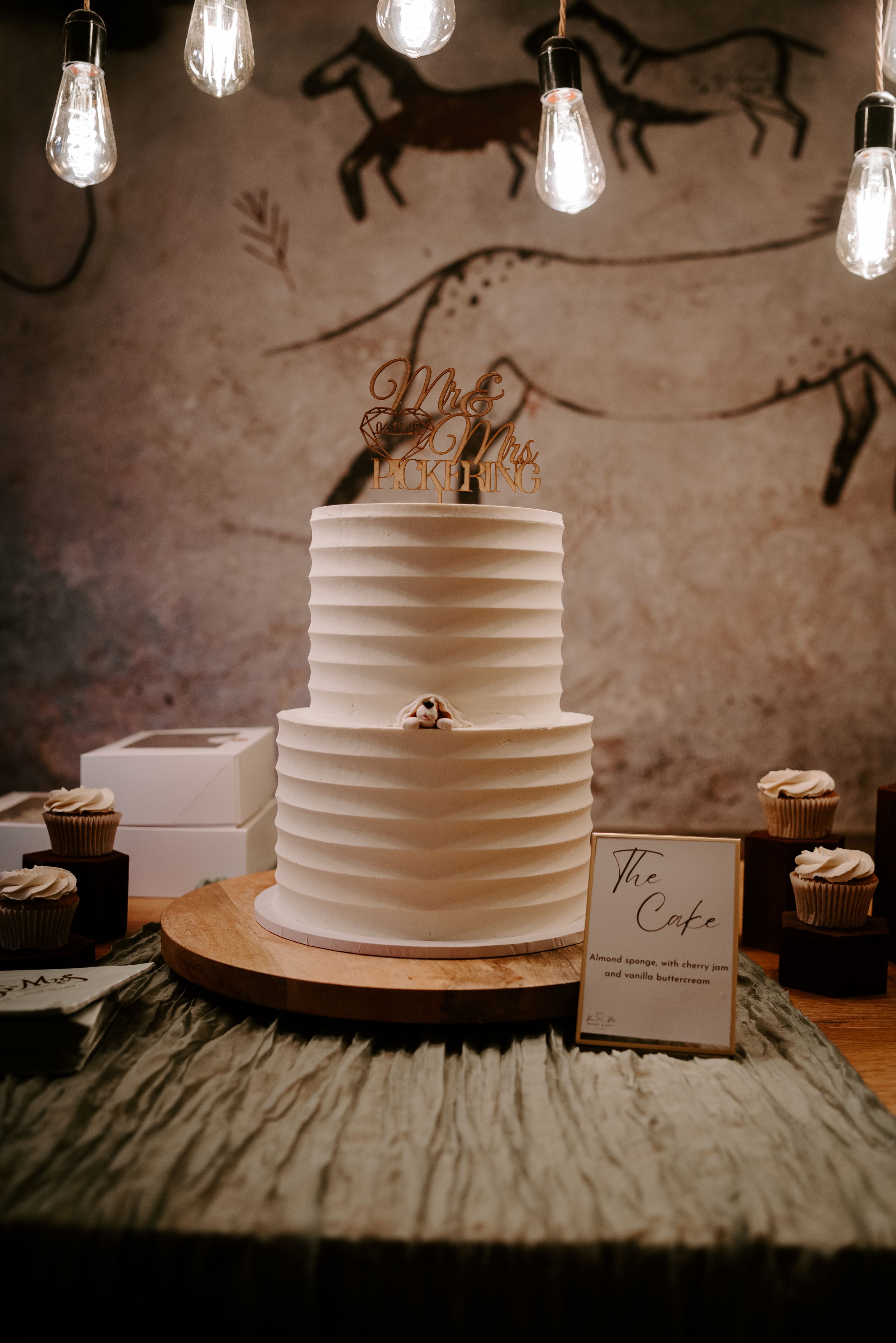 A series of simple white wedding cakes with horizontal lines displayed on wooden stands under hanging lights.