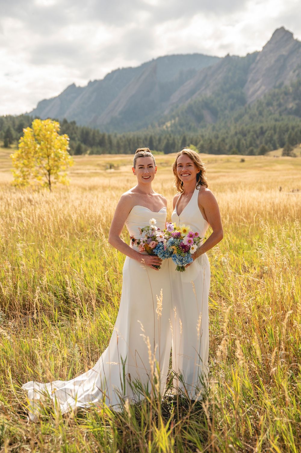 Two people in white dresses hold colorful bouquets while standing in a golden field with mountain peaks in the background.