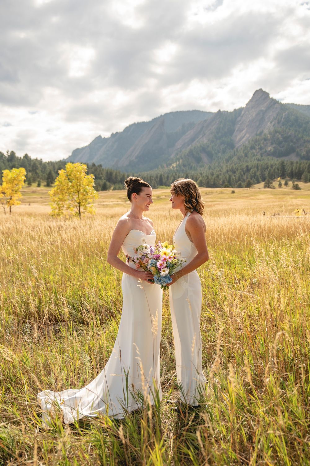 Two people in white dresses hold bouquets while standing in a golden field with mountain peaks in the background.