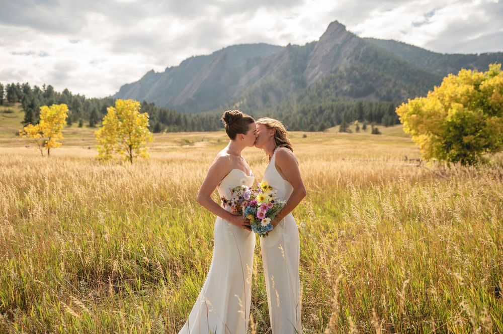 Couple in white dresses share intimate moment in mountain meadow with dramatic cloudy sky and autumn colors.