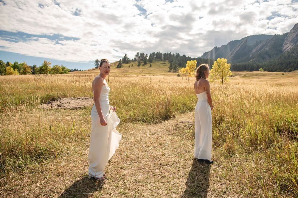Two people in white dresses walk through a golden field at sunset with mountains in the background.