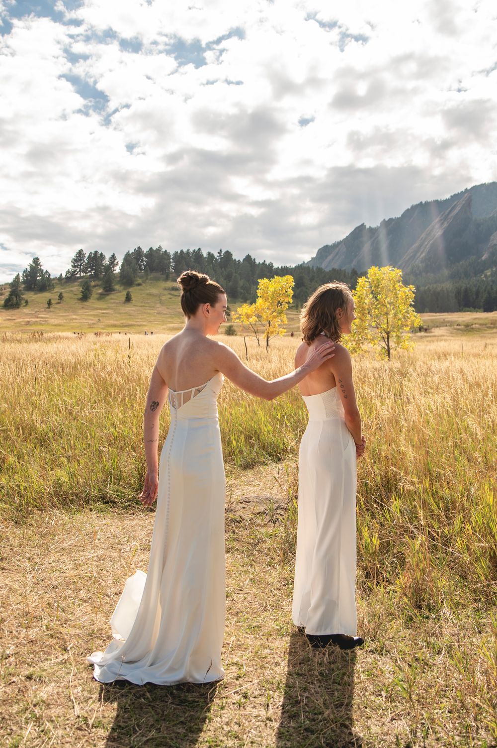 Two figures in white dresses stand together in a golden field with mountains and trees in the background at sunset.