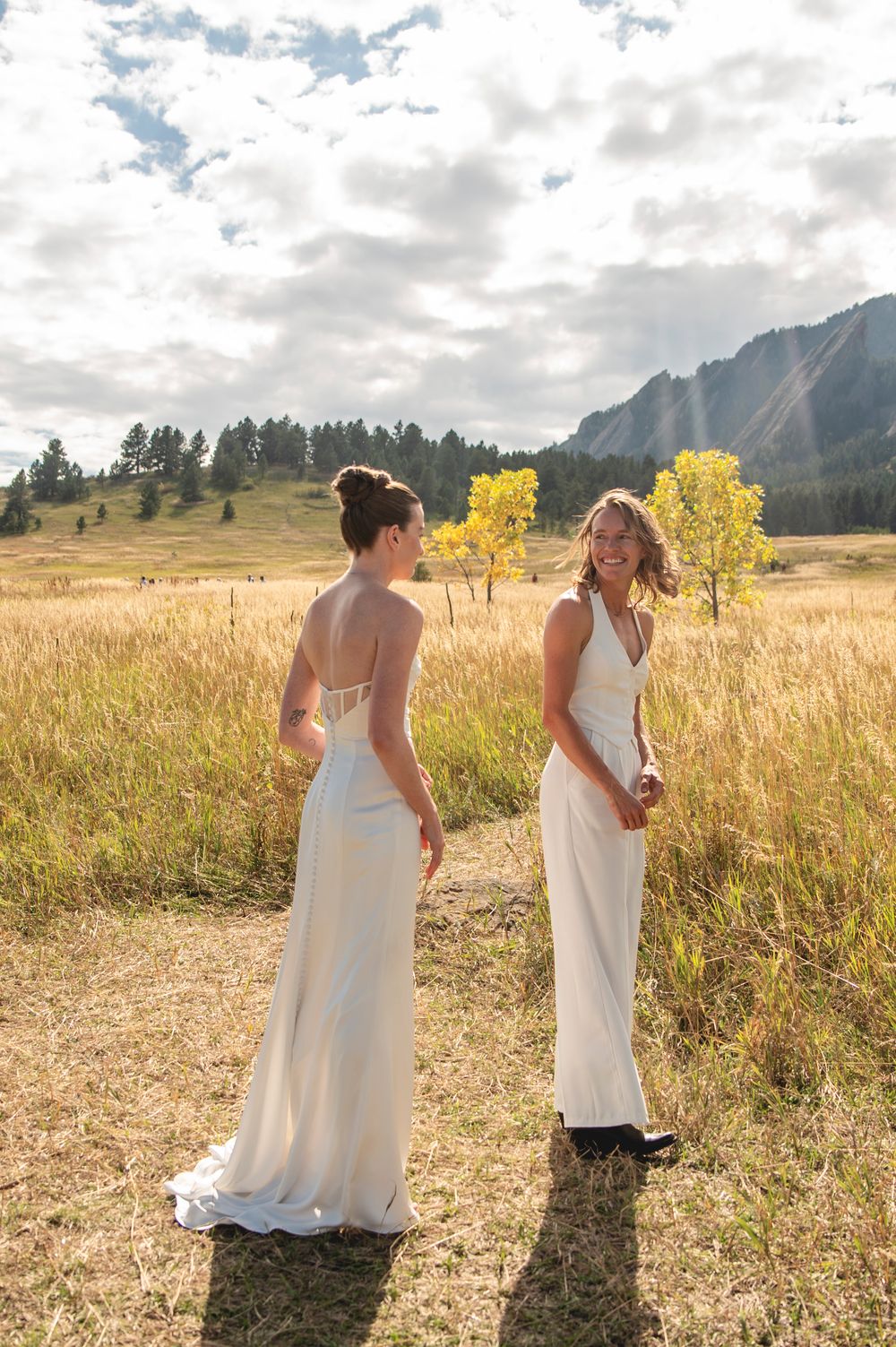 Two people in white dresses walk through a golden meadow with mountain views and autumn foliage.
