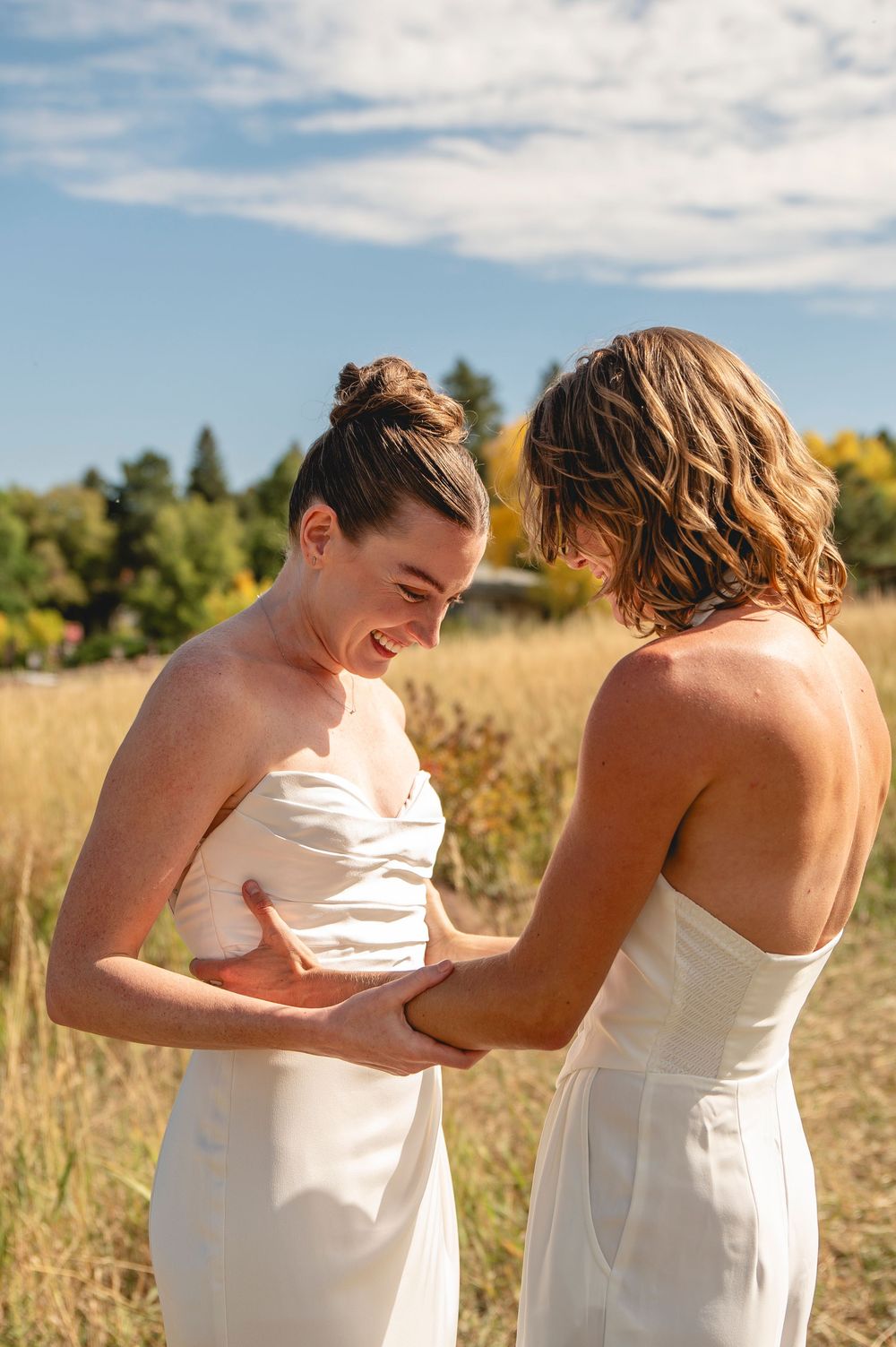 Two people in white dresses share a joyful moment together in a sunlit field with golden grass and blue sky.