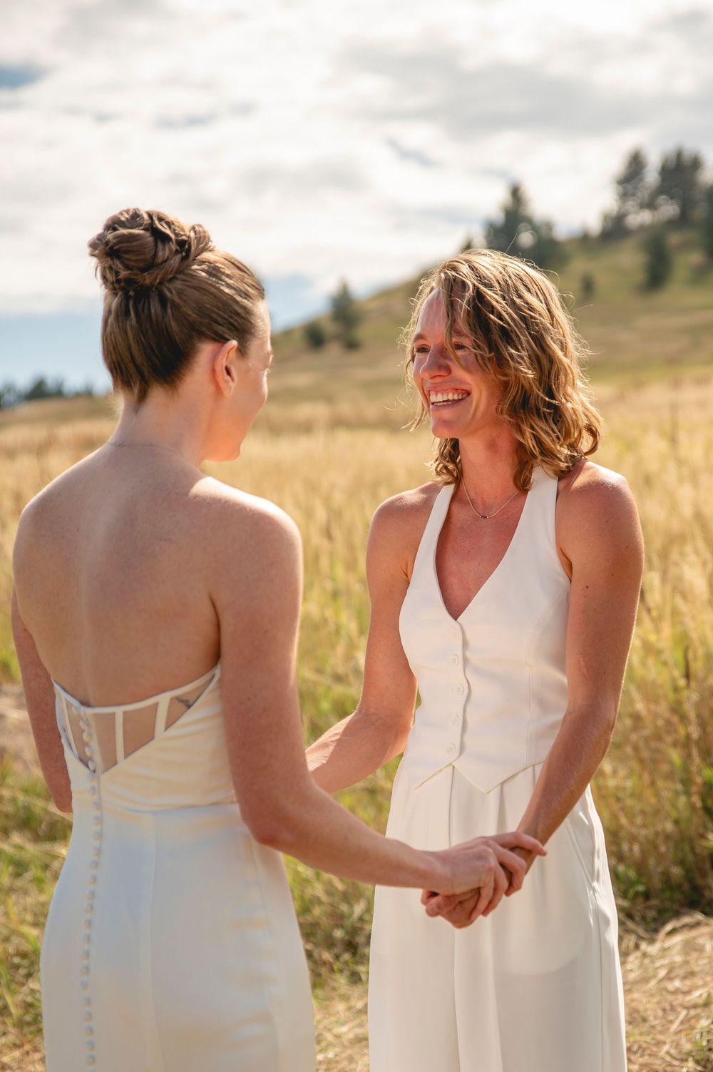 Two people in white dresses hold hands while standing in a golden field during sunset.