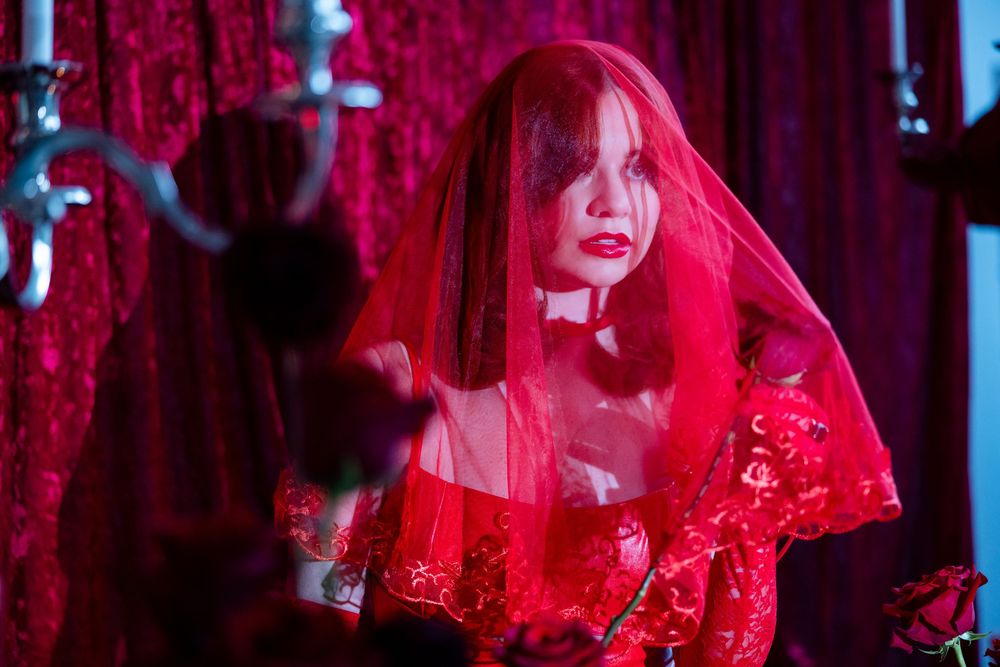 Woman in red lace veil and off-shoulder gown posing against a red velvet backdrop.