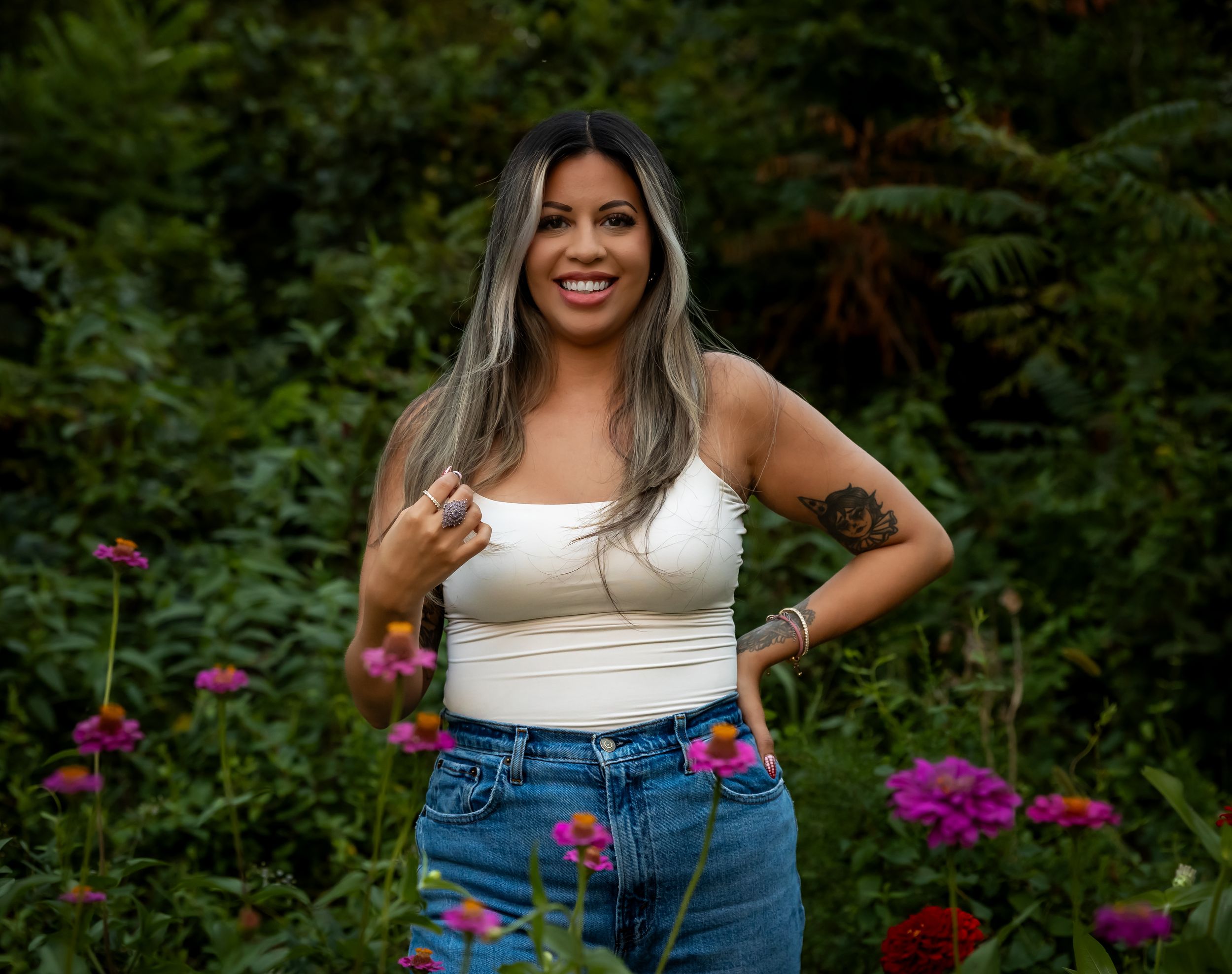 Woman in casual attire poses in garden with vibrant purple flowers in background.