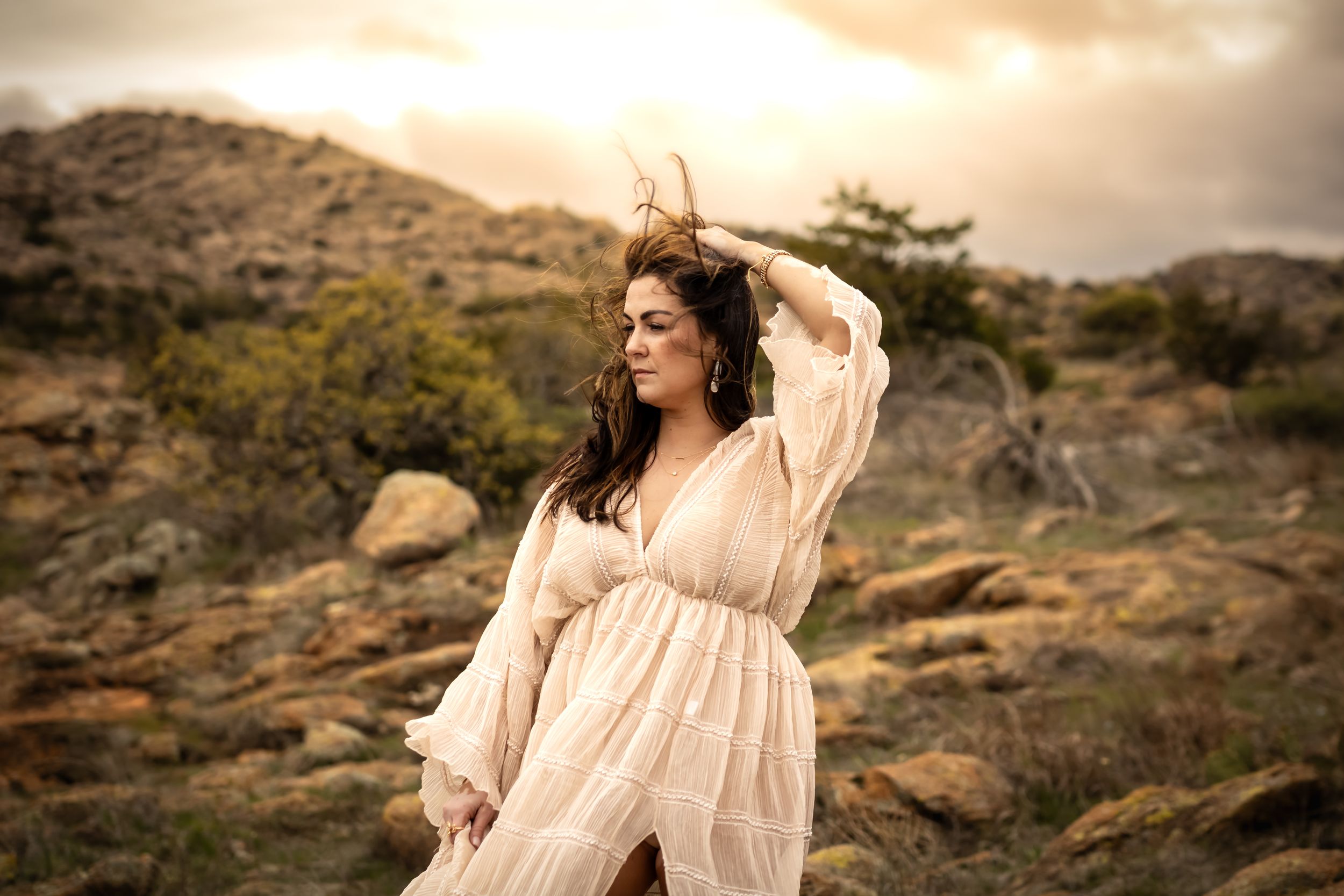 Woman in flowing cream dress poses on rocky terrain at golden hour.