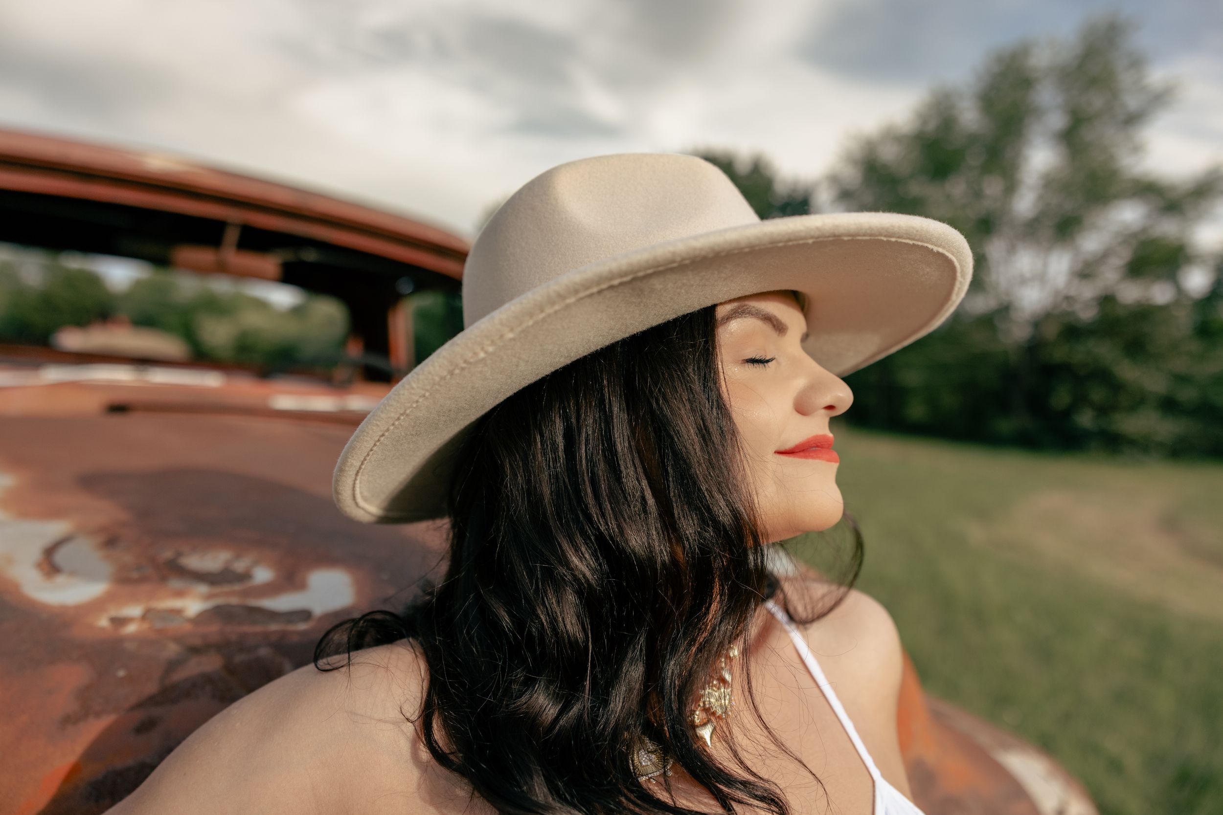 Woman wearing wide-brim hat poses against rustic outdoor setting.