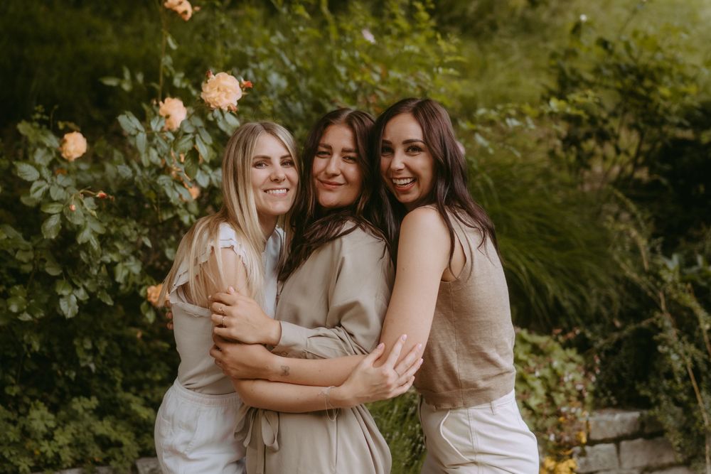 Friends in neutral tones embracing and smiling in a garden with blooming roses.