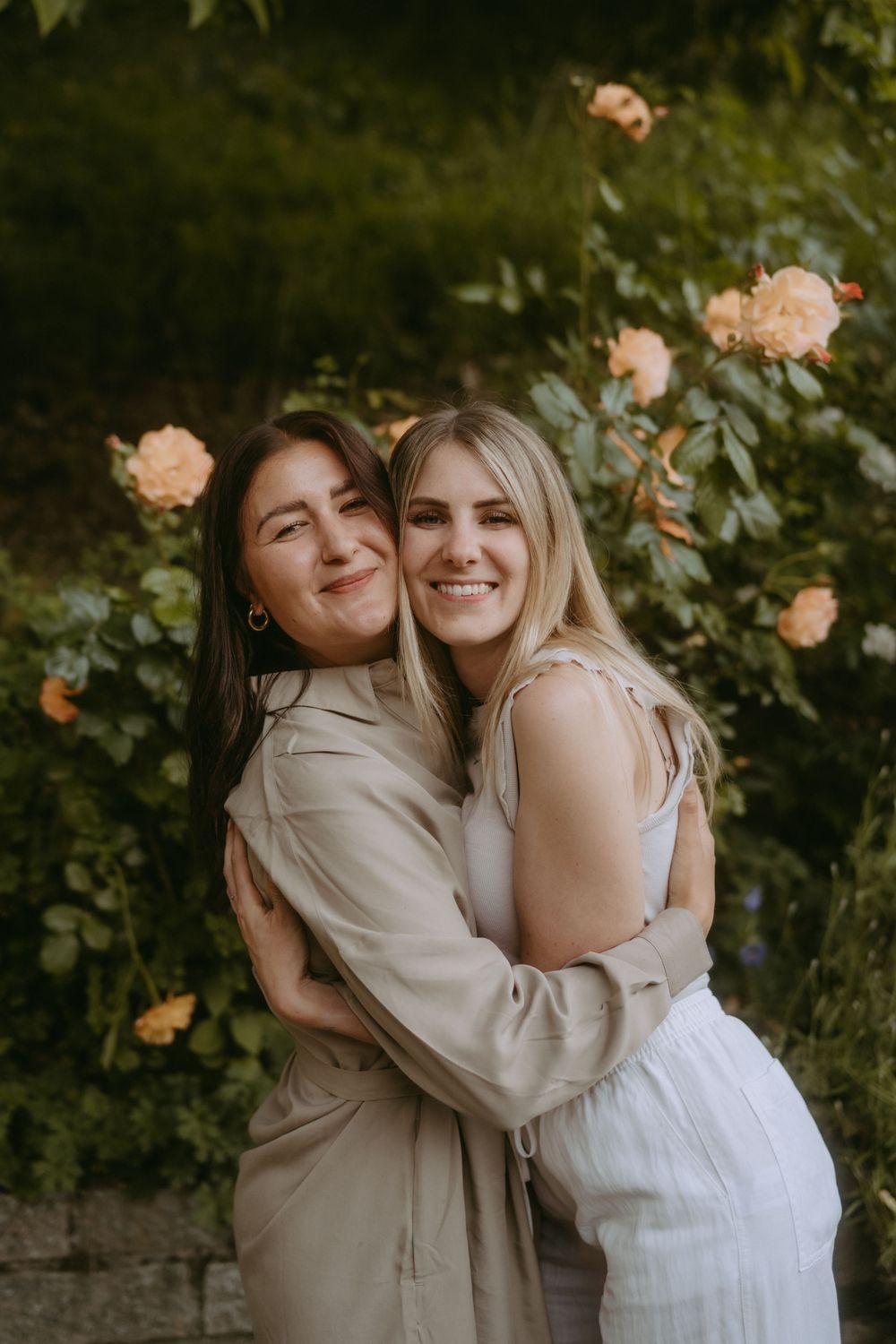 Candid moment of friends hugging and laughing in a garden with peach roses.