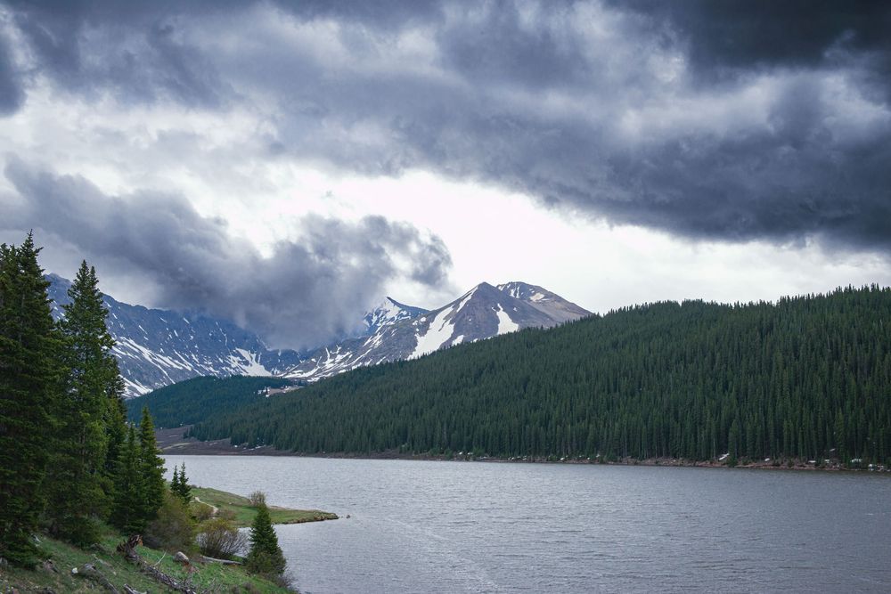 Dramatic storm clouds gather over snow-capped Colorado mountain peaks and evergreen forests reflected in a pristine alpine lake.