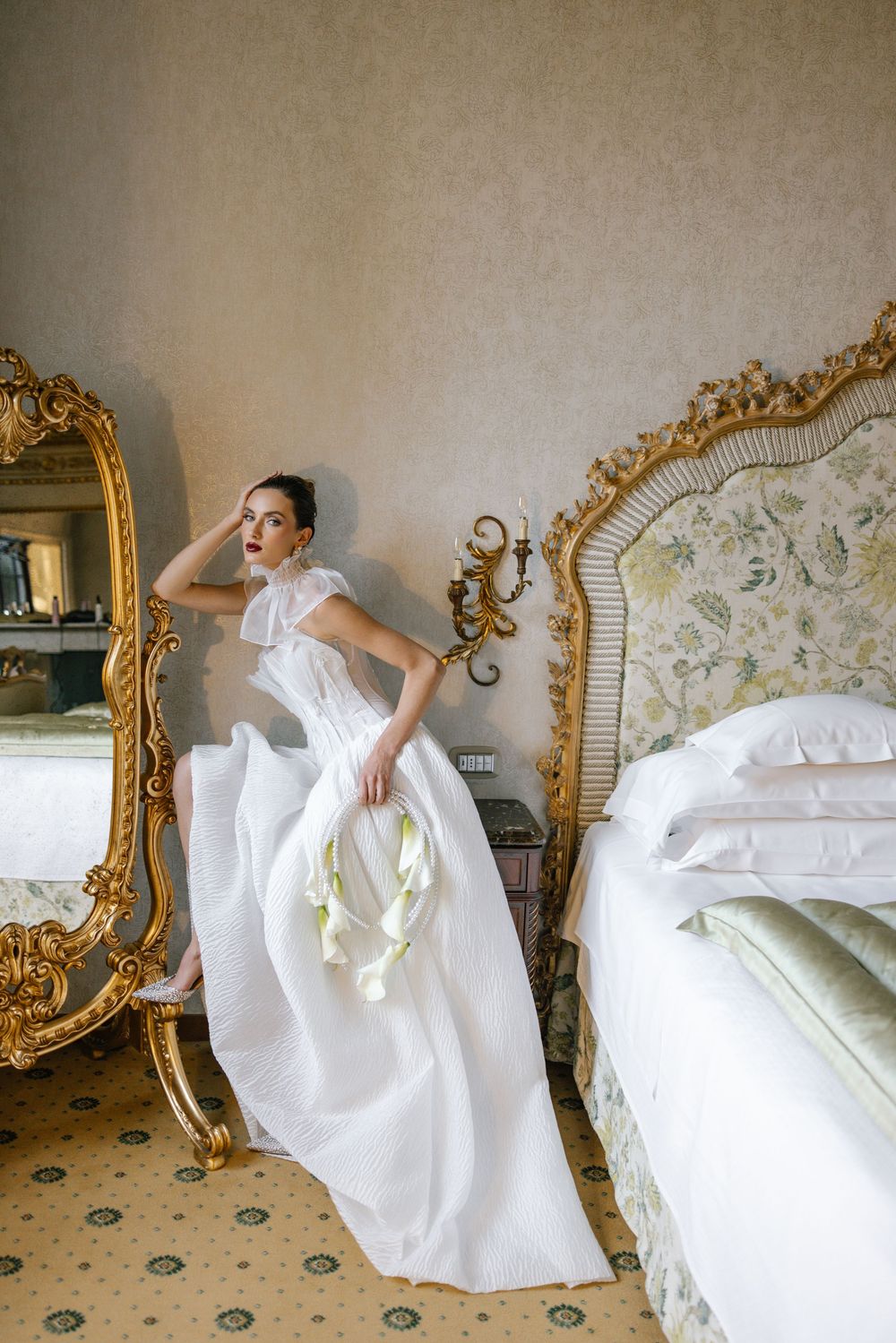 Elegant bridal fashion photo in ornate bedroom with gold mirrors and vintage decor featuring flowing white gown.