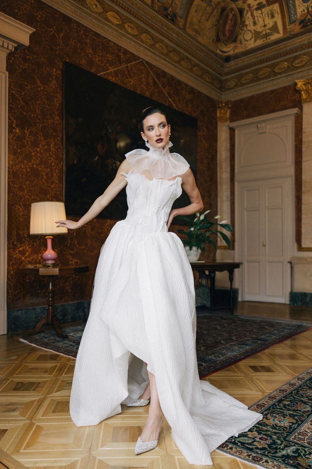 Dramatic bridal gown with high neck and full skirt photographed in luxurious vintage room setting.