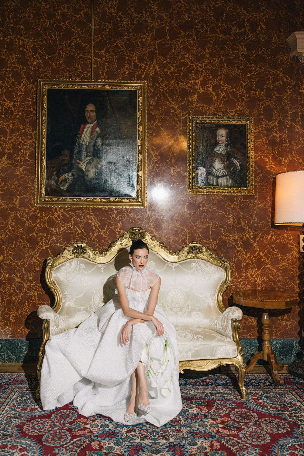 Bride in white dress reclining on vintage sofa against ornate wallpaper with framed portraits.