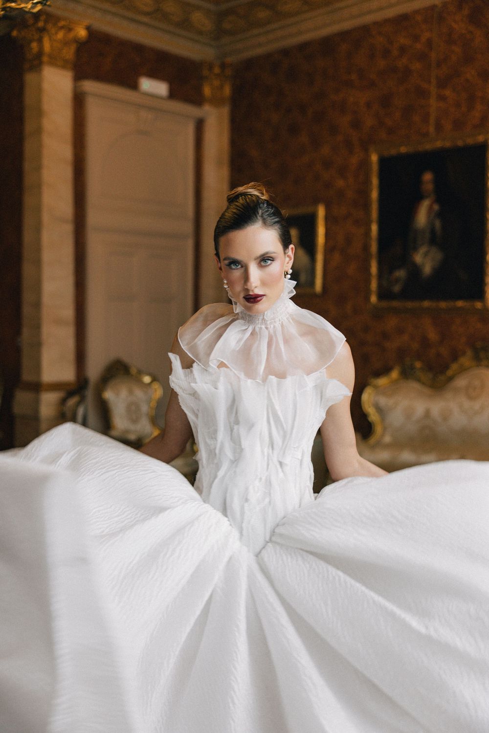 Dramatic wedding gown with high ruffled collar and voluminous skirt photographed in ornate vintage room.