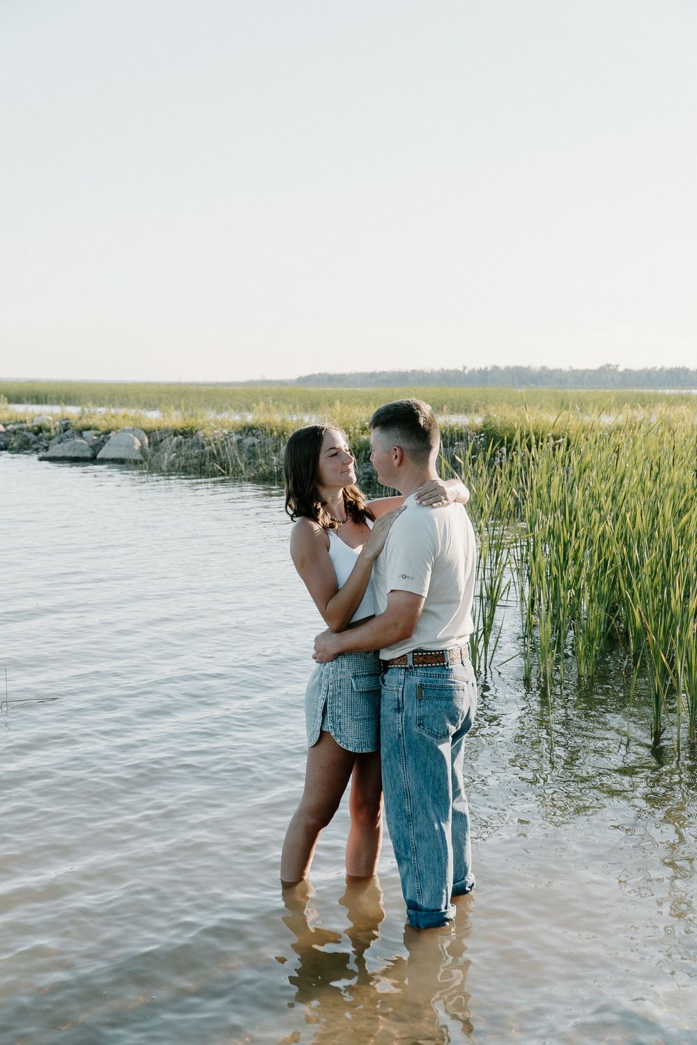 A romantic couple stands together in shallow water near reeds during a summer photoshoot at sunset.