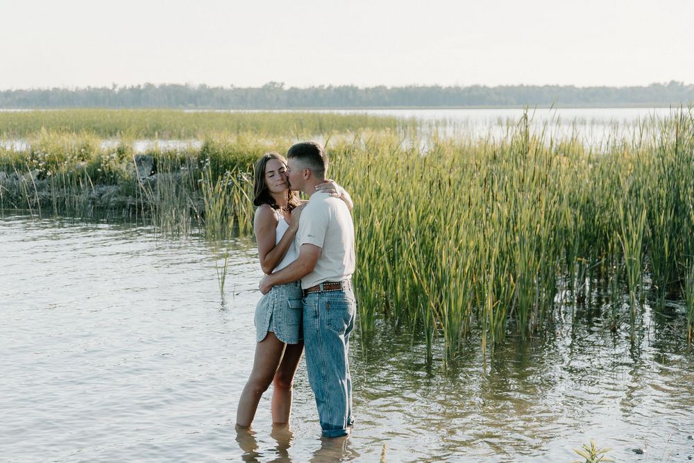 A couple shares an intimate moment while standing in shallow water surrounded by tall reeds at sunset.