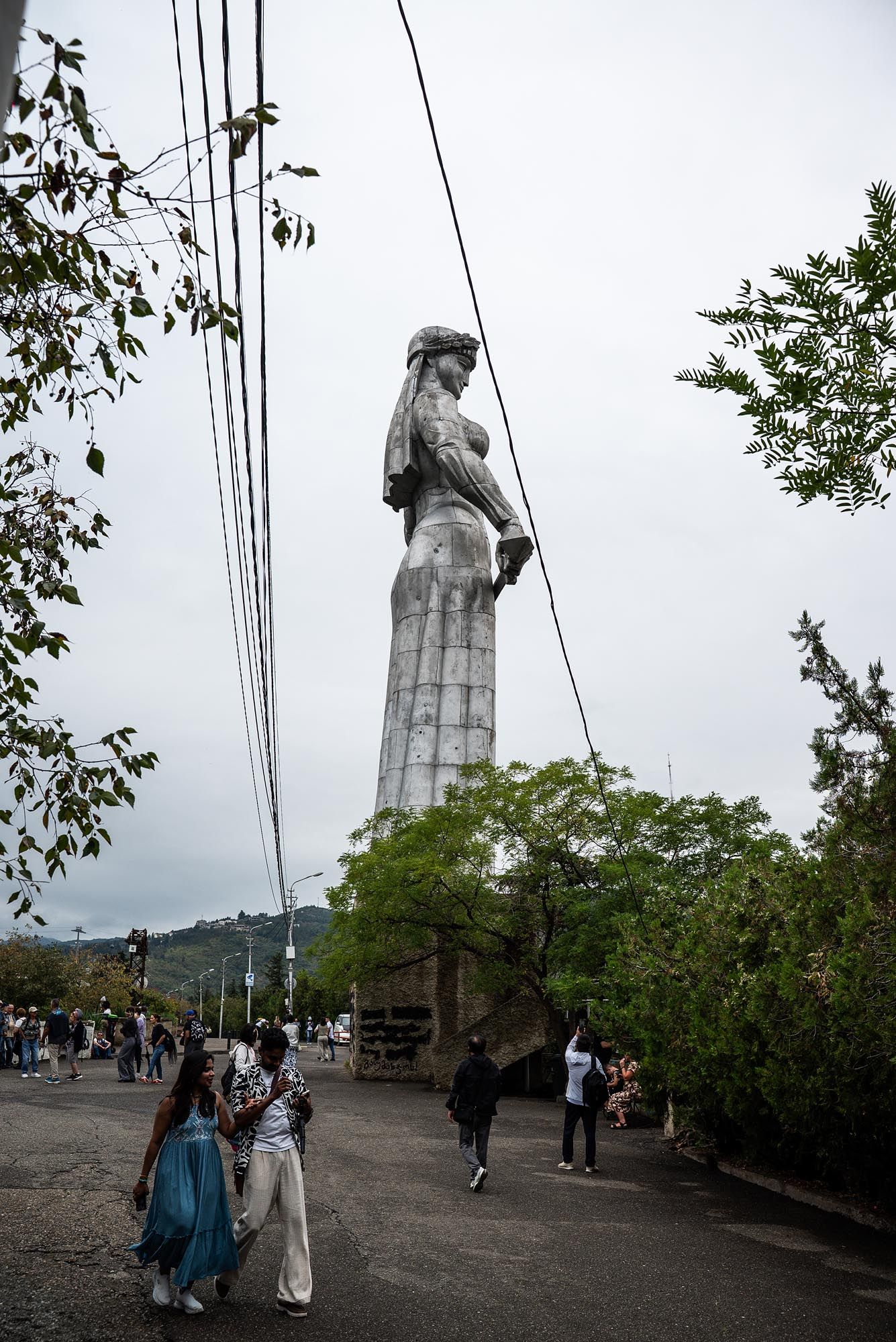 Tall stone monument statue stands against cloudy sky with power lines and trees in Tbilisi, Georgia.