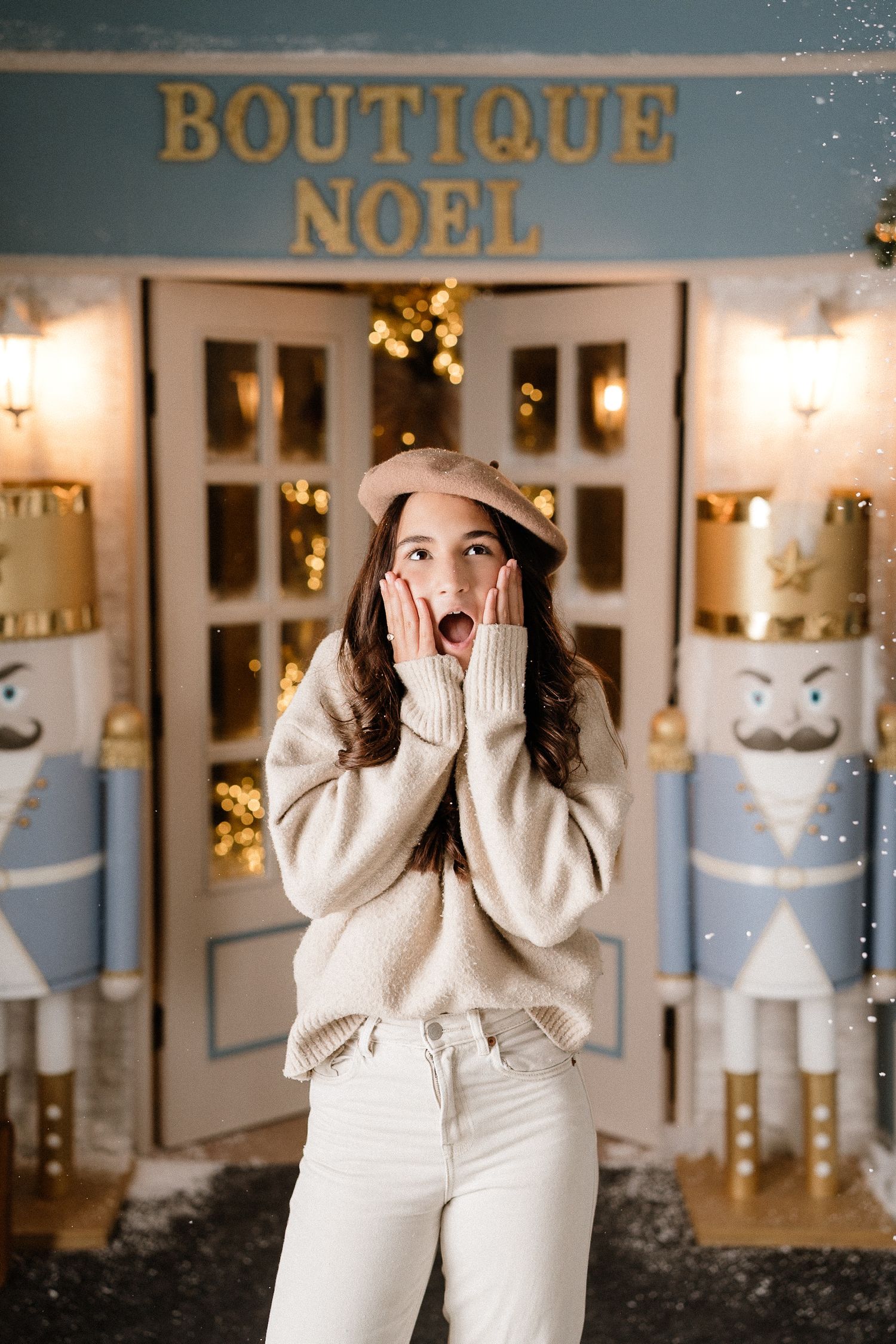 A smiling baby in a white sweater sits in a white high chair with Christmas decorations and lights in the background.