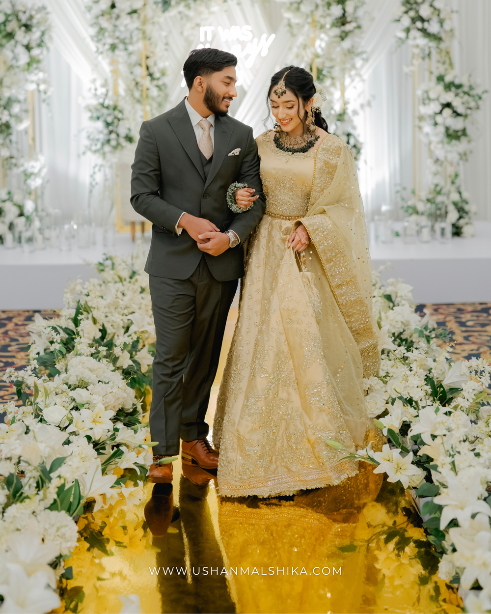A couple in traditional wedding attire pose amid white floral decorations and a golden walkway.