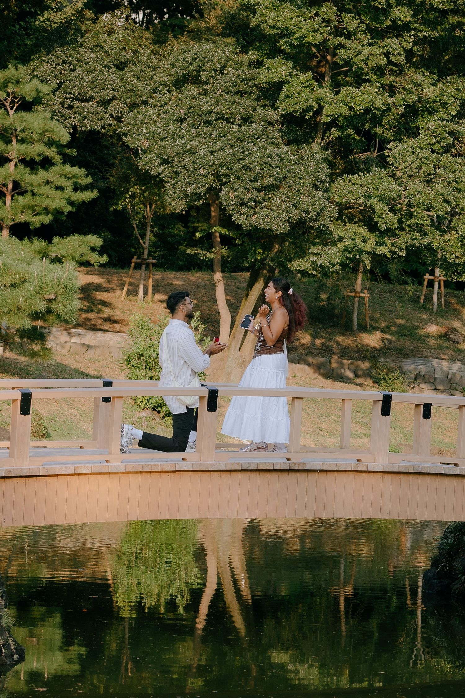 A romantic proposal moment on a wooden bridge over a reflective pond surrounded by lush green trees.