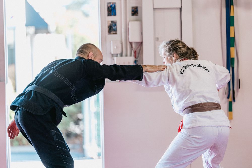 Students practicing martial arts moves with partners in a bright training studio with white walls and blue mats.