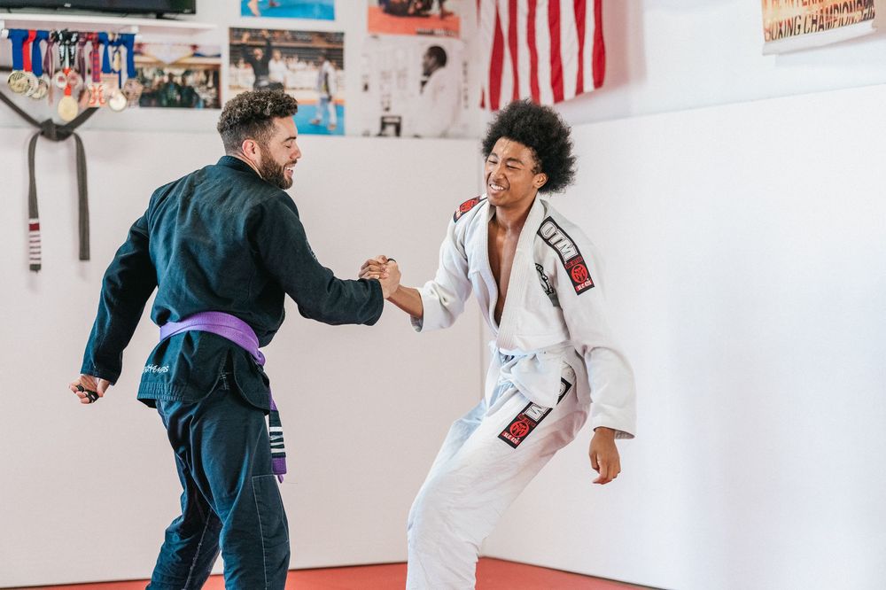 Two martial artists practicing Brazilian Jiu-Jitsu techniques on training mats in a gym with an American flag on the wall.