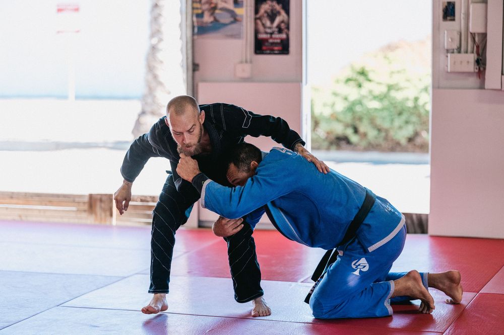 Two martial artists practicing grappling techniques on a red training mat in a bright studio space.