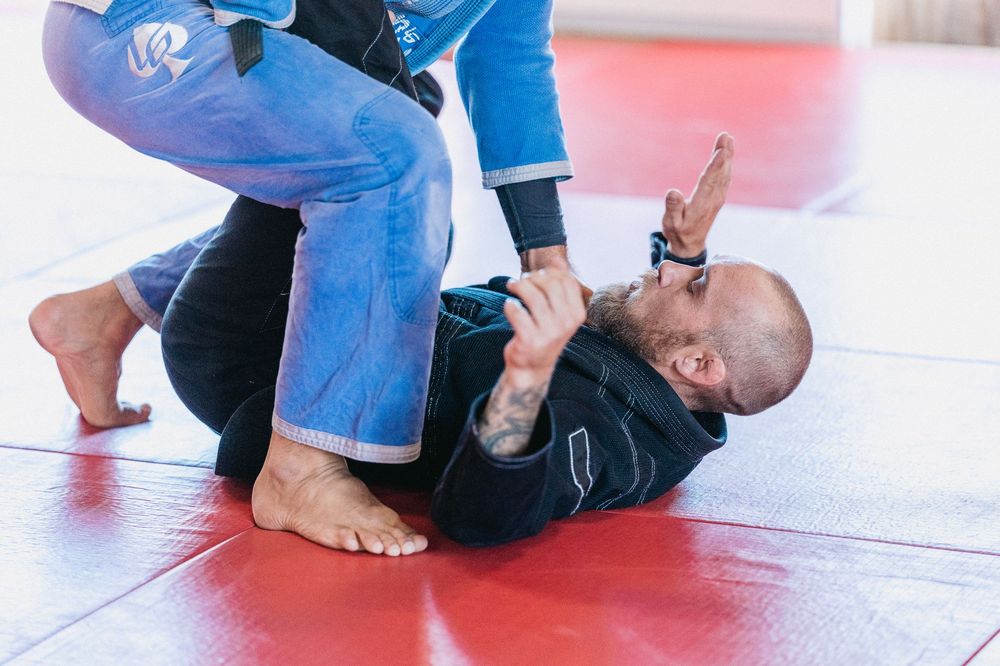 Two martial artists practicing Brazilian Jiu-Jitsu on red mats, with one attempting a submission from guard position.