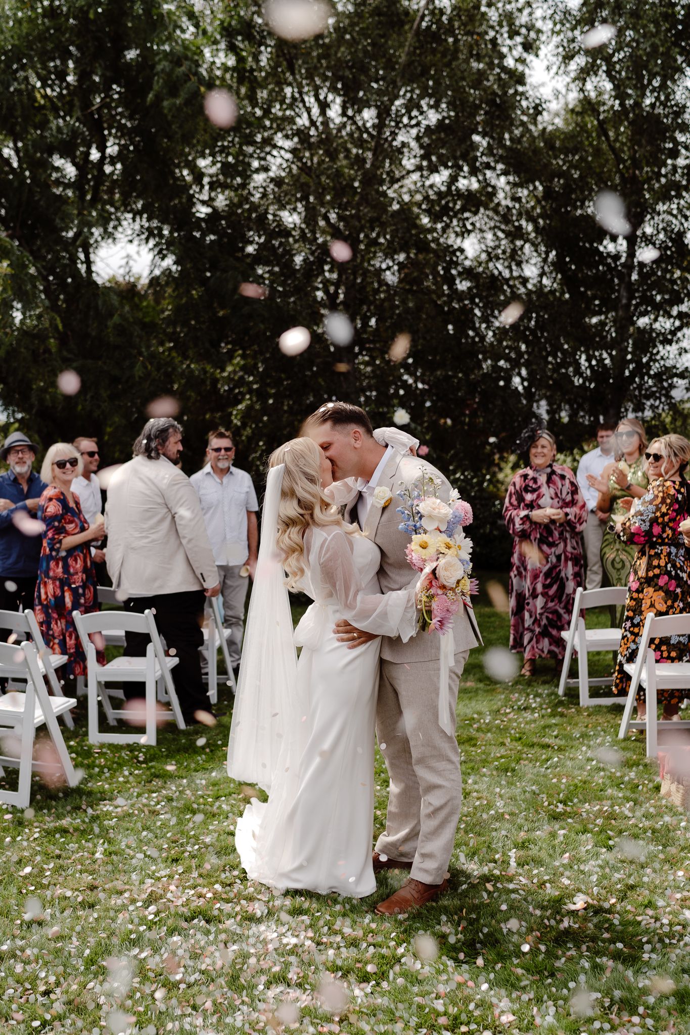 A modern natural photo of a wedding couple kissing as they exit the ceremony with confetti from The Whole Bride showering down on them