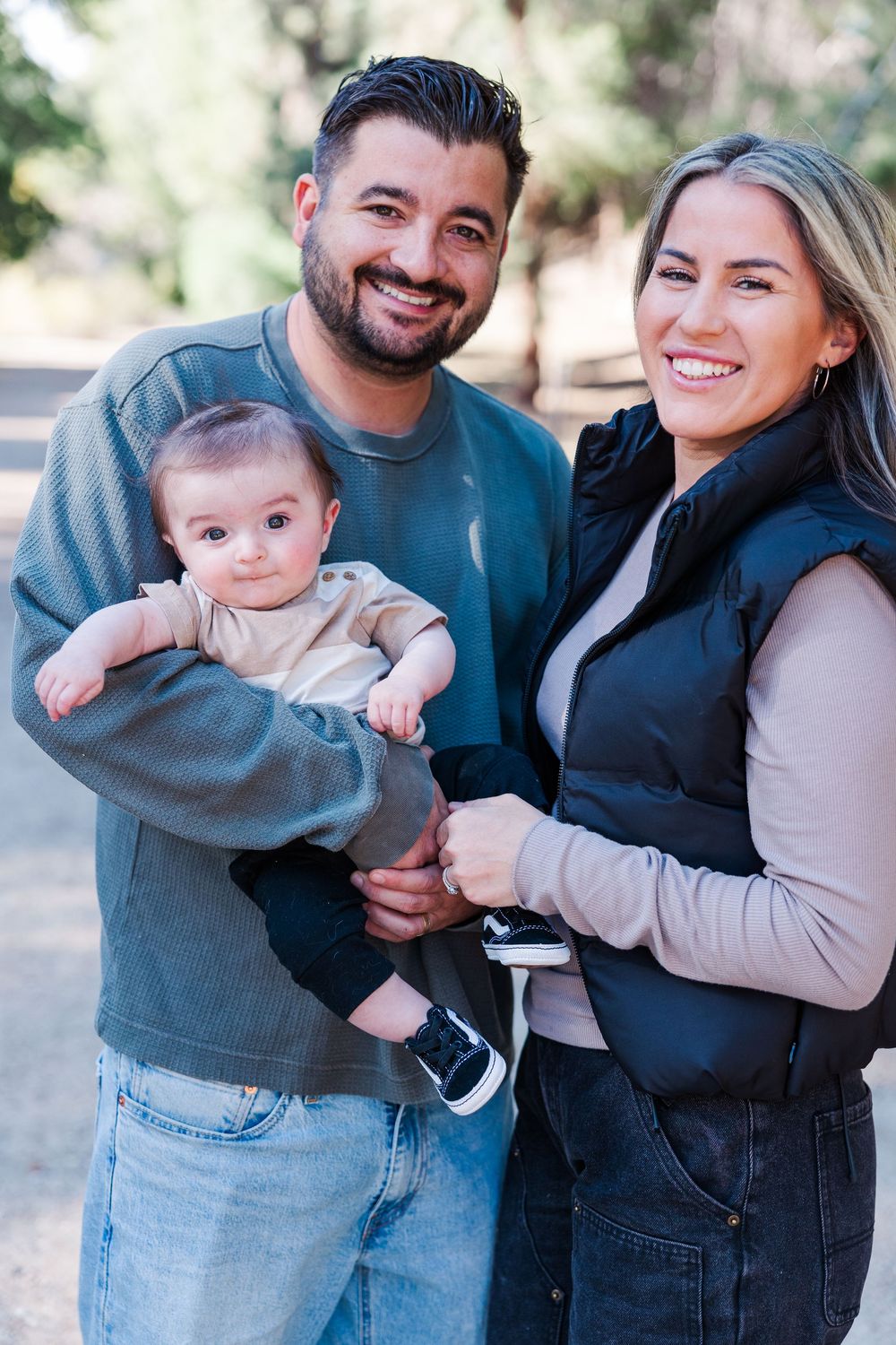 A happy family of three poses together outdoors on a sunny day wearing casual clothing.