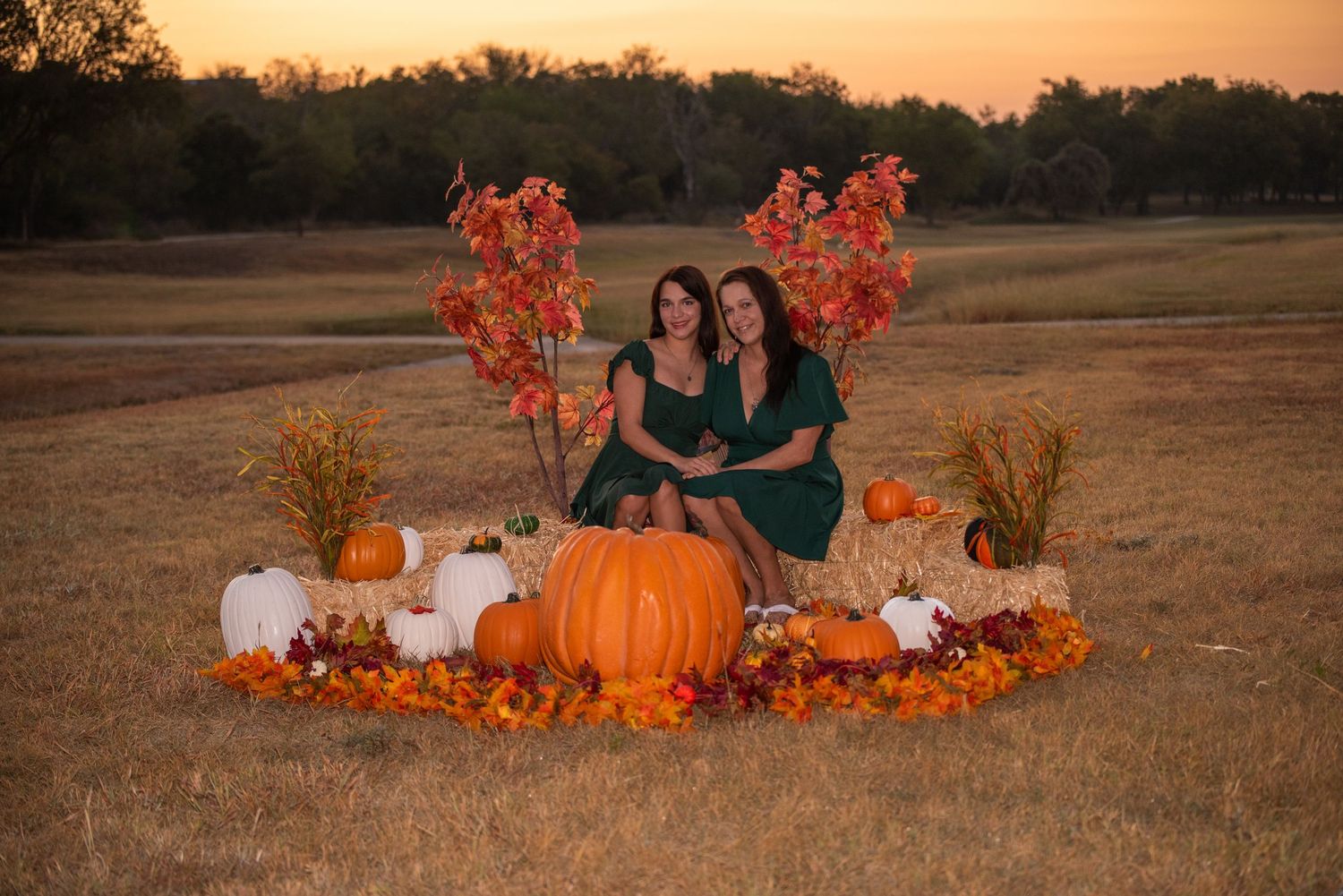 Autumn sunset photoshoot setup with orange pumpkins, fall foliage and white decorative elements in a rustic outdoor field.