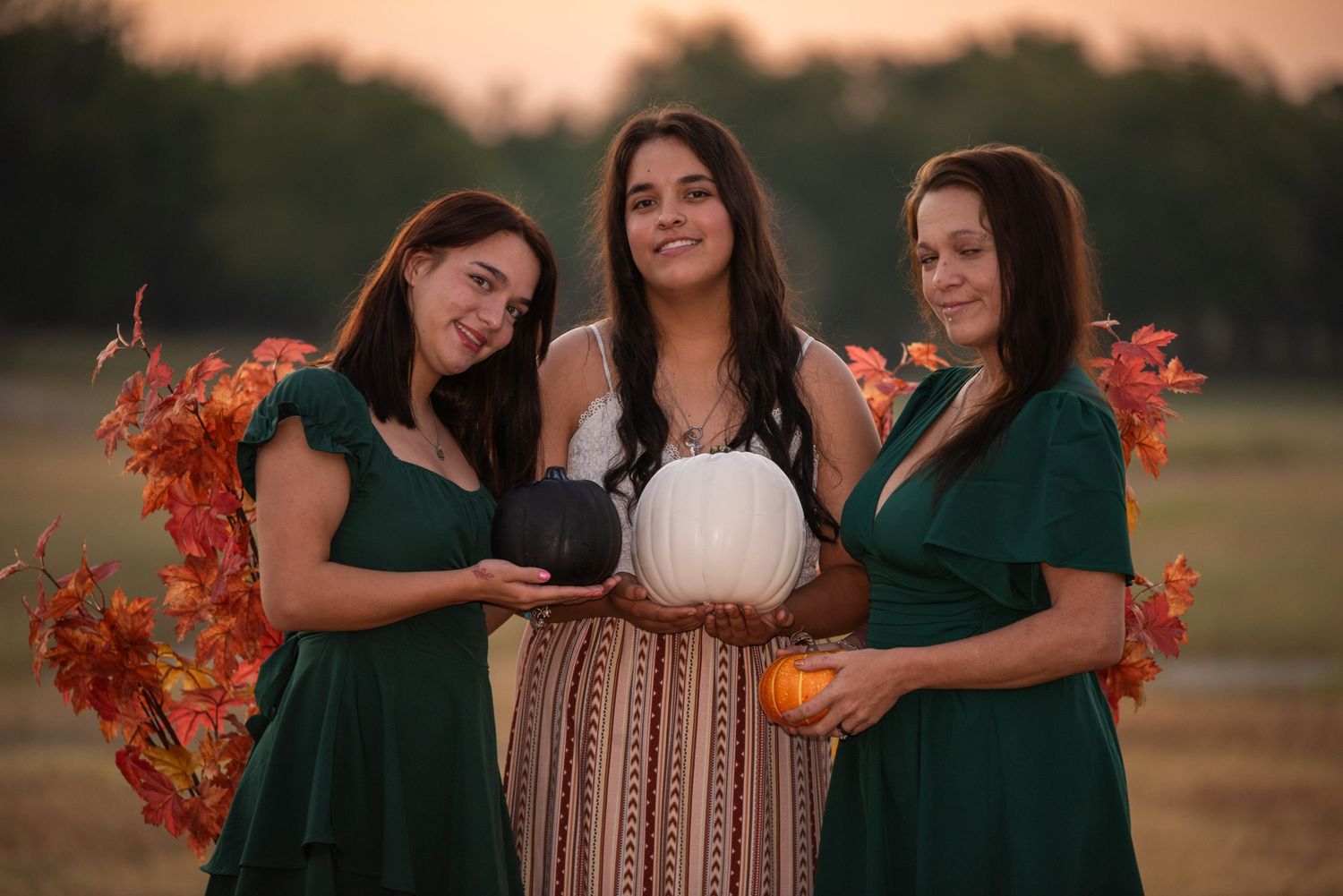 Three women in coordinating green and white dresses pose together during sunset in a fall outdoor setting.