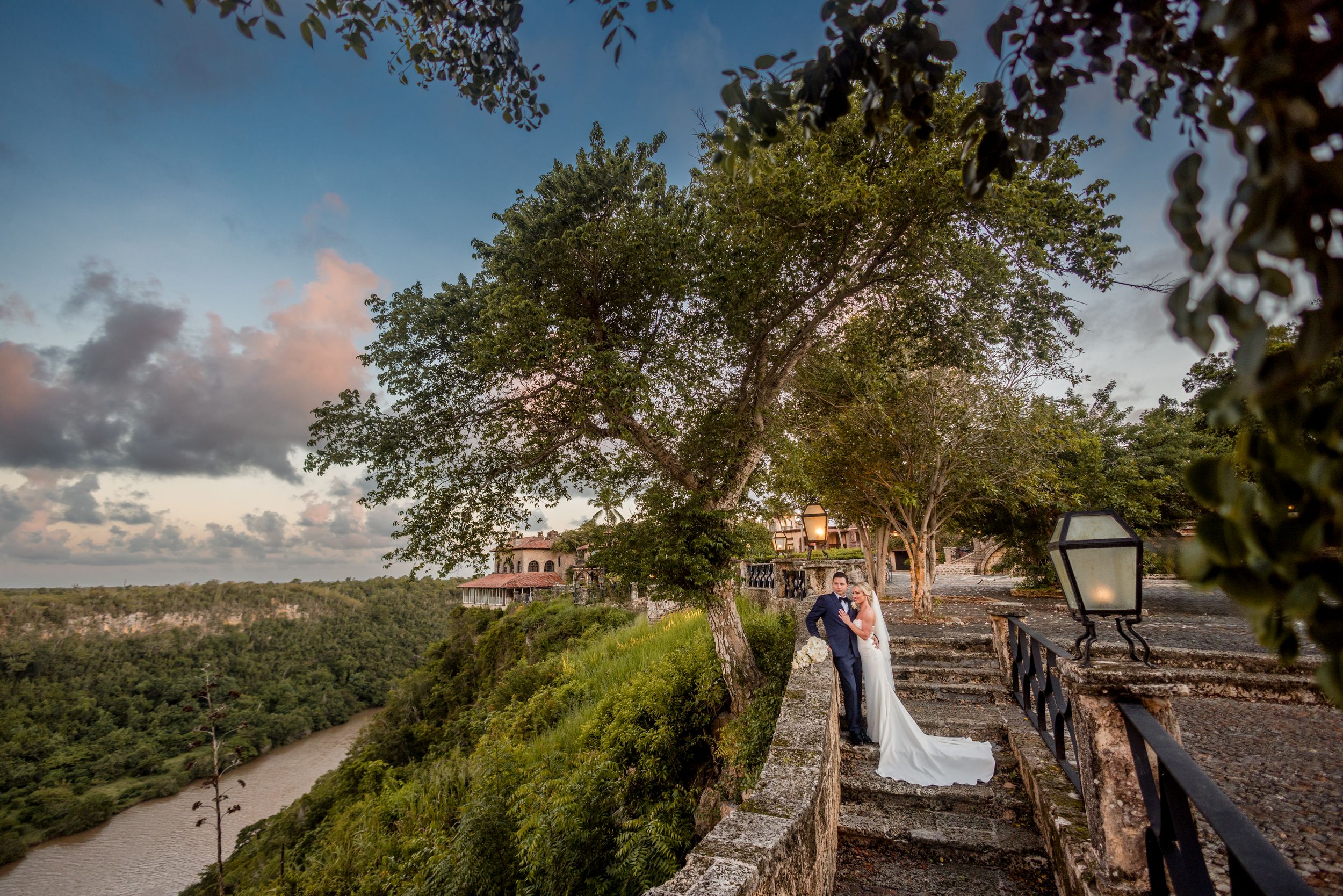 Scenic coastal boardwalk with wooden railings extends along the shoreline as clouds drift across the blue sky at sunset. Altos de Chavon