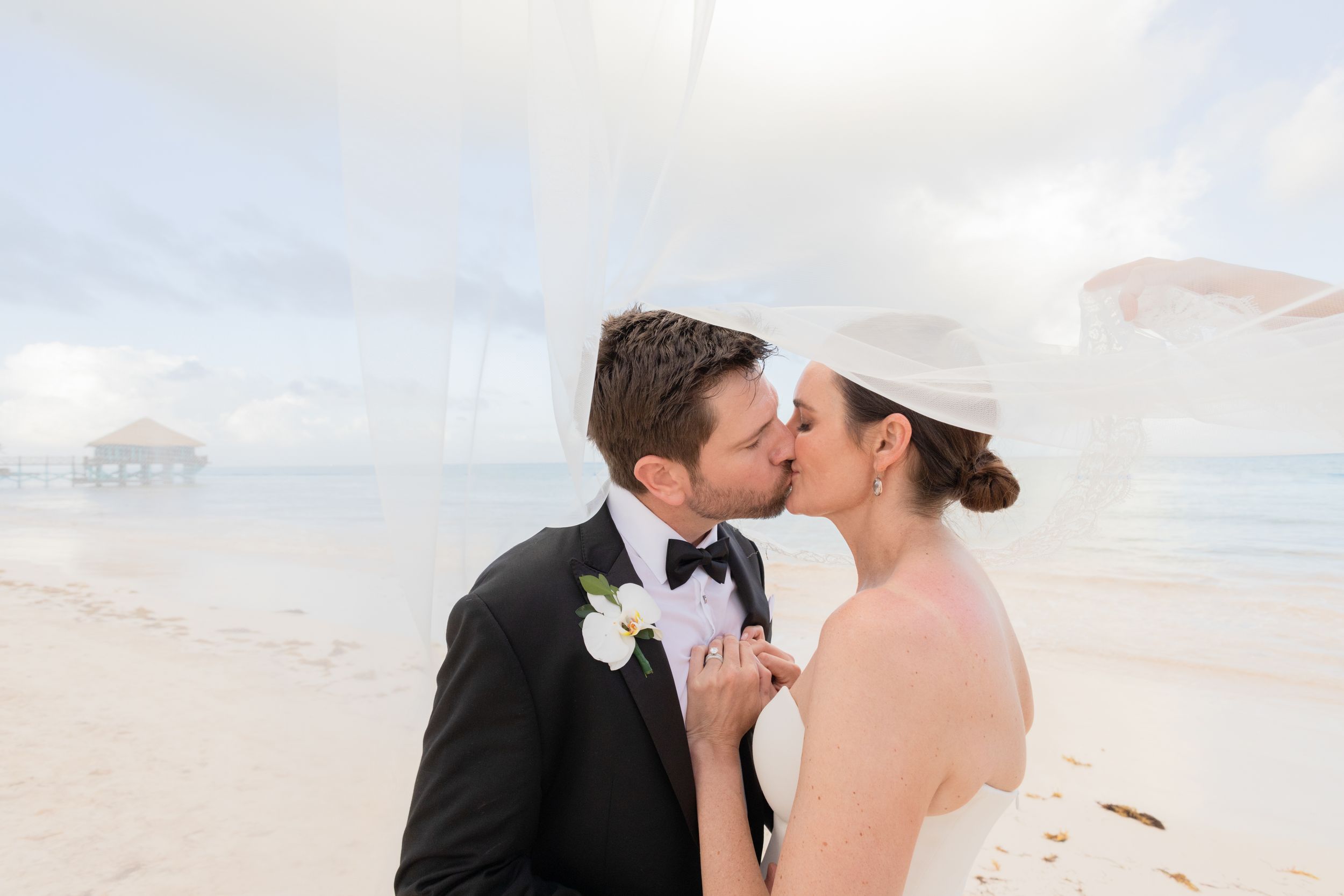 A couple shares a romantic kiss on a beach at sunset with soft clouds and ocean waves in the background.