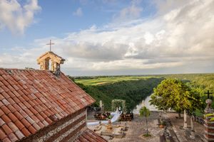 Panoramic view of a historic church with red tiled roof overlooking a lush green valley under cloudy skies.
