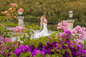 Bride and groom embracing on a stone terrace surrounded by vibrant pink and purple flowers with lush greenery.
