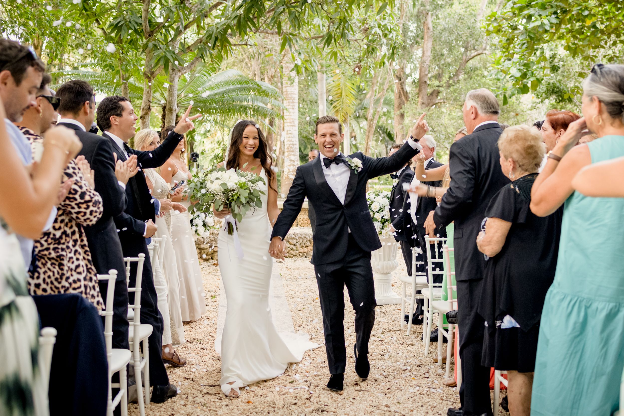 Wedding party celebrates with joyful reactions during outdoor ceremony in a tree-lined setting.