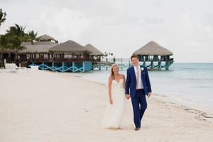 A couple walks along a tropical beach with overwater bungalows and turquoise ocean waters in the Maldives. Sanctuary Cap Cana