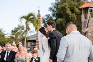 A romantic wedding ceremony moment at an outdoor venue with palm trees and stone architecture in the background.