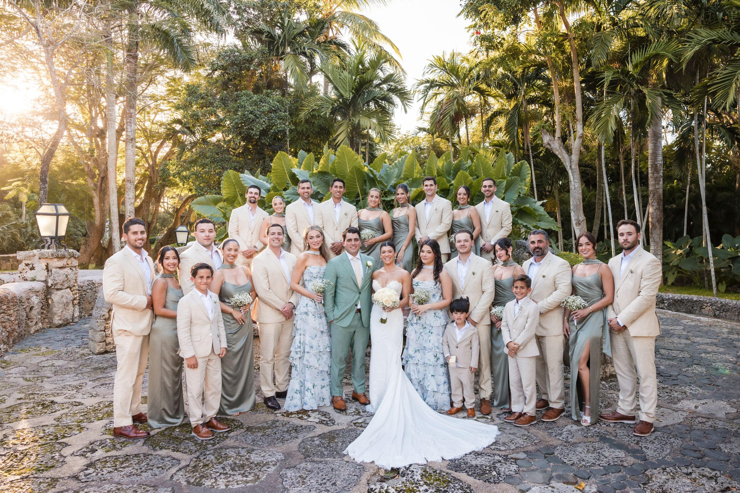 Wedding party poses in neutral and sage green attire against a tropical garden backdrop during sunset.